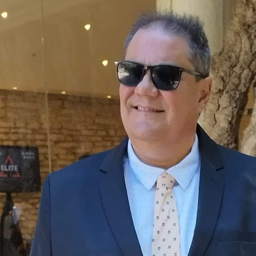 Foto do autor: Homem de pele clara, cabelo curto grisalho, usando fato clássico azul escuro, camisa social clara e gravata bege com quadradinhos, além de óculos escuros, sorrindo em ambiente bem iluminado.